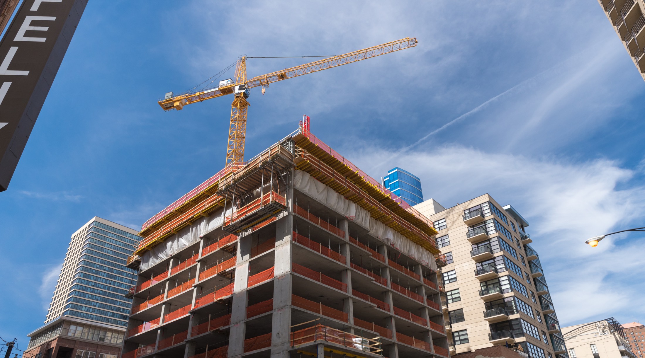 A mid-rise building under construction, with a tower crane above it and completed towers alongside.