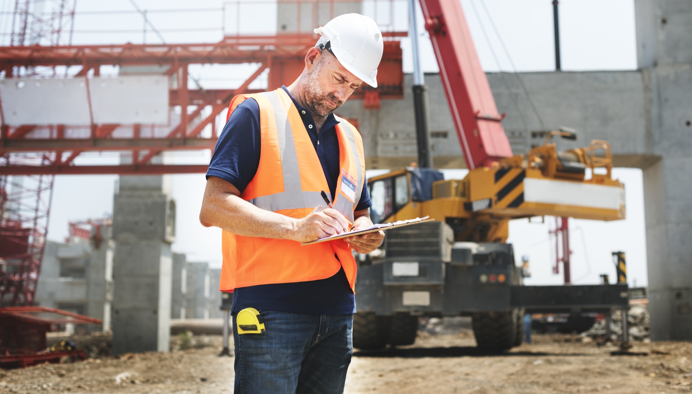 A site supervisor in hi-vis making notes on a clipboard, with a crane and part-built structure behind.